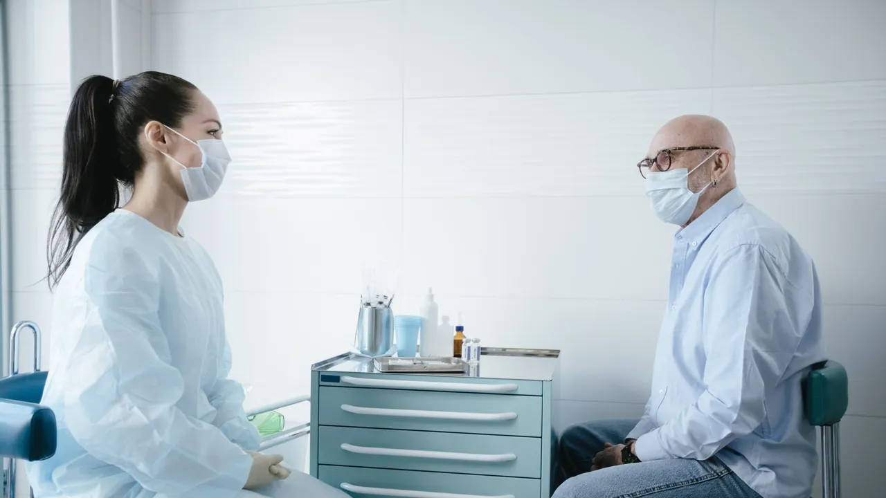 A medical professional wearing PPE checks a patient's pulse indoors.